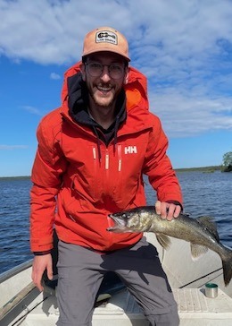 Mathieu wearing a red coat, standing on a boat, and holding a fish