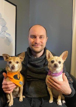 Patrick en chandail gris et pantalon gris dans son salon, tenant ses chihuahuas Mika et Abby